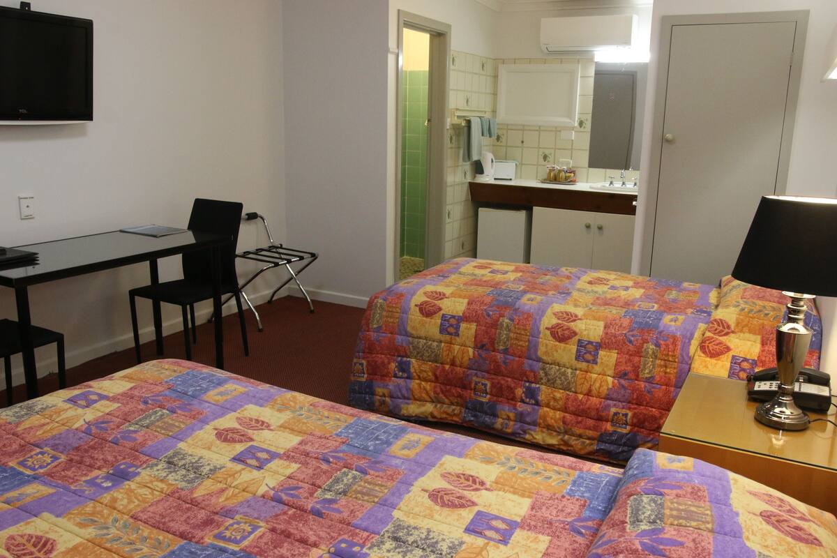 Habitación de hotel con dos camas, un escritorio, una silla, un televisor, una lámpara y un baño visible a través de una puerta abierta.