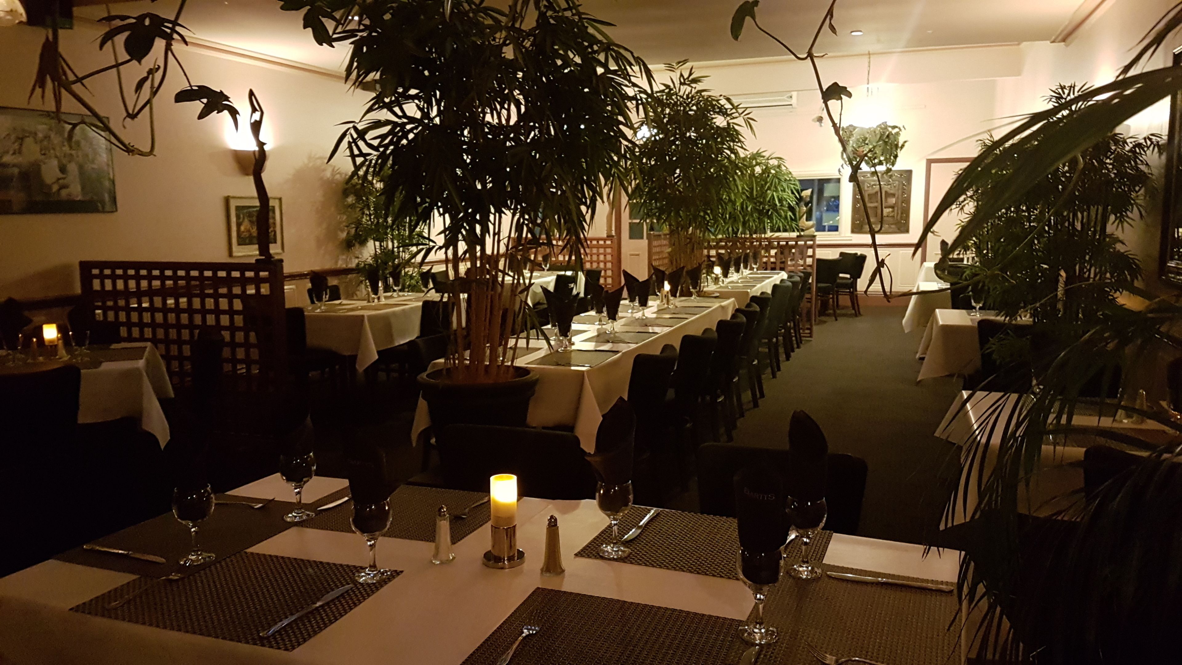 A dimly lit restaurant with tables set for dining, featuring a warm ambiance and indoor plants.