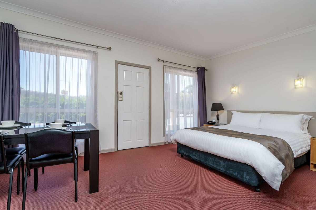 Habitación de hotel con una cama, un escritorio con sillas, una mesa de comedor y dos ventanas con cortinas.