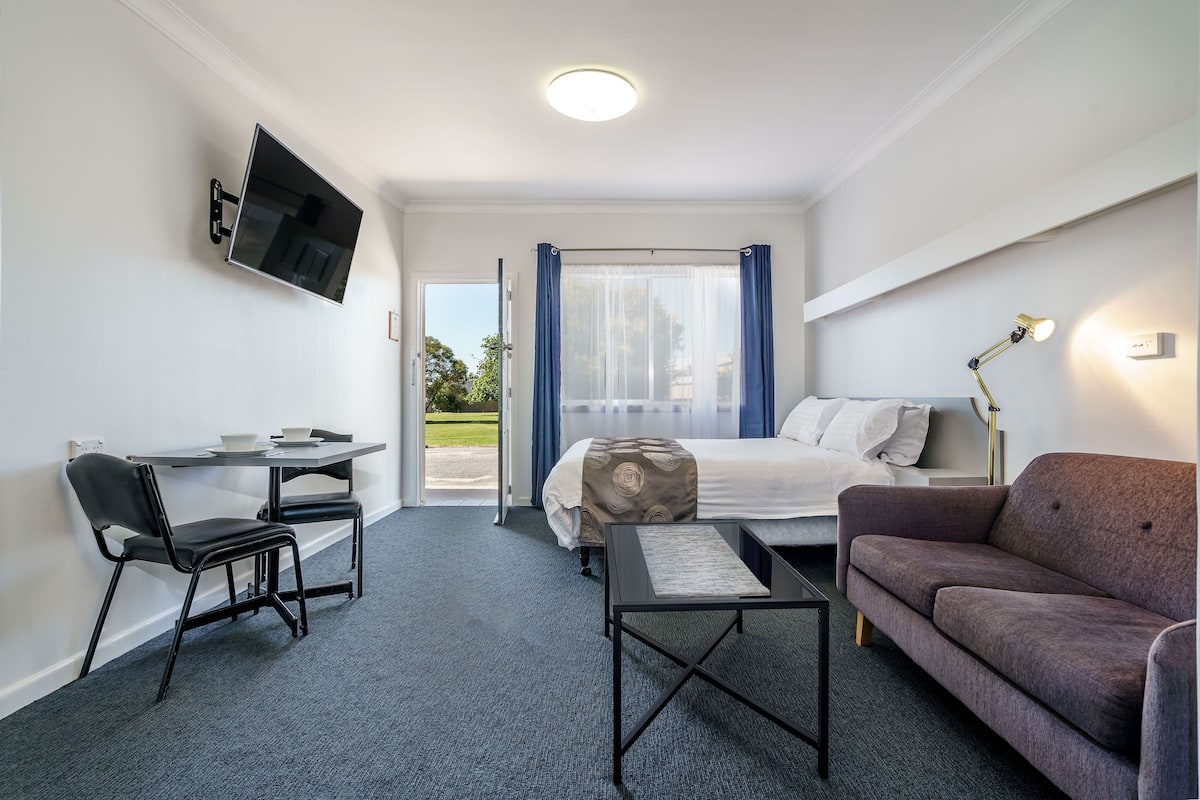 Habitación de hotel con cama, escritorio con silla, sofá, televisor montado en la pared y ventana con cortinas.