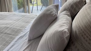 A bedroom with a large bed, multiple pillows, and a view of a green landscape through a sliding glass door. - Langbrook Estate Cottages (Yarra Junction)