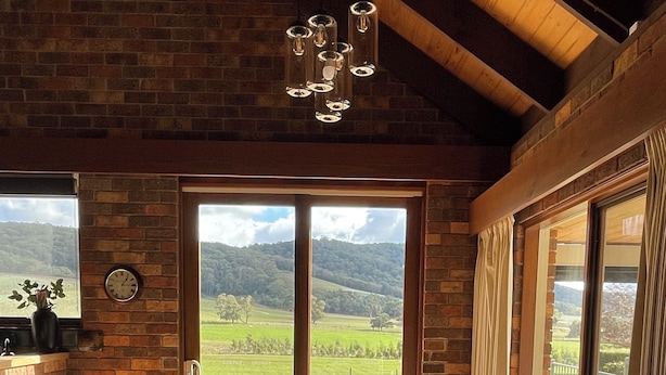 A dining area with a wooden table and chairs, a hanging light fixture, and a view of a green landscape through large windows. - Langbrook Estate Cottages (Yarra Junction)