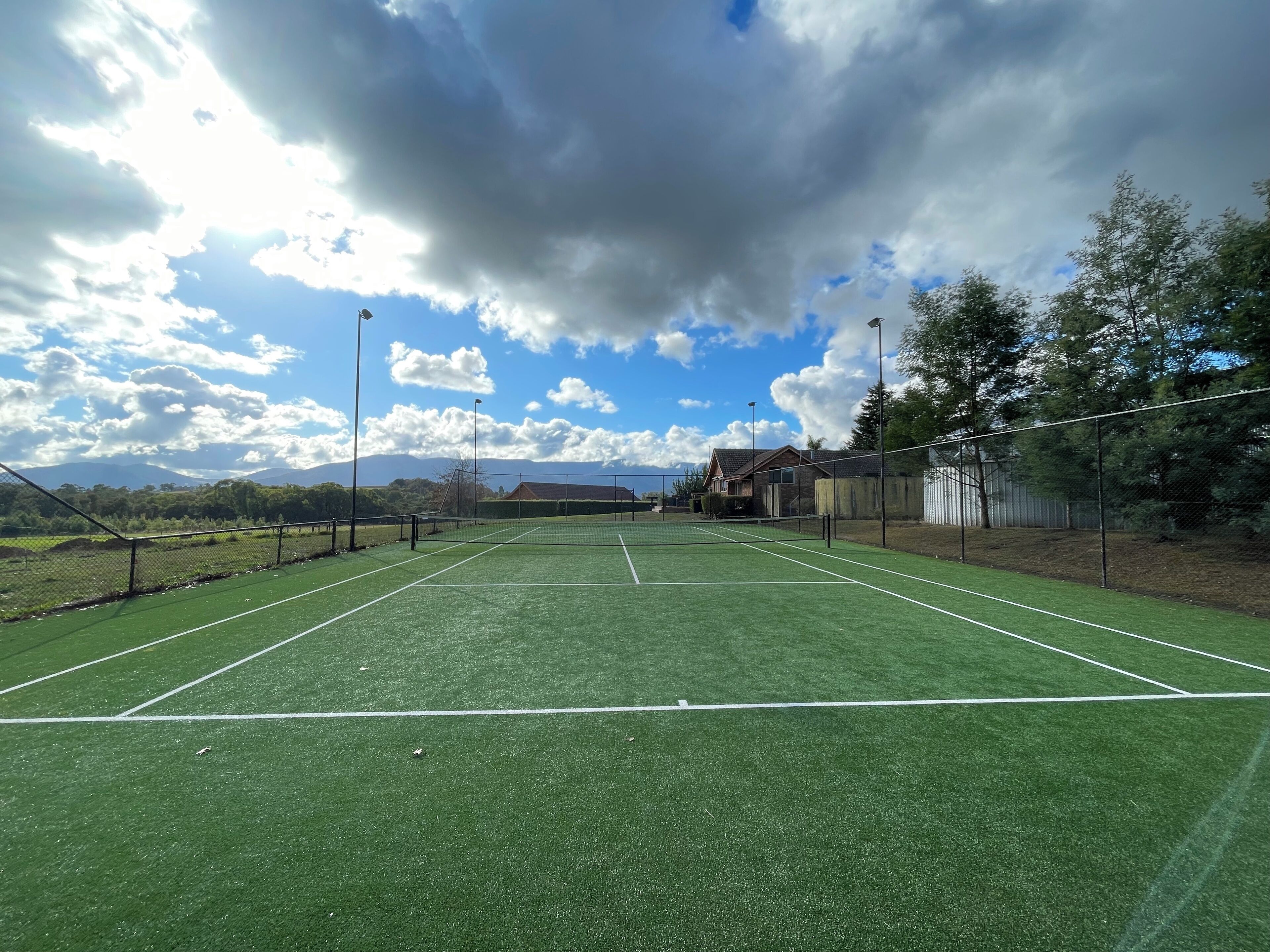 An outdoor tennis court with a net, surrounded by a fence, and a house in the background.