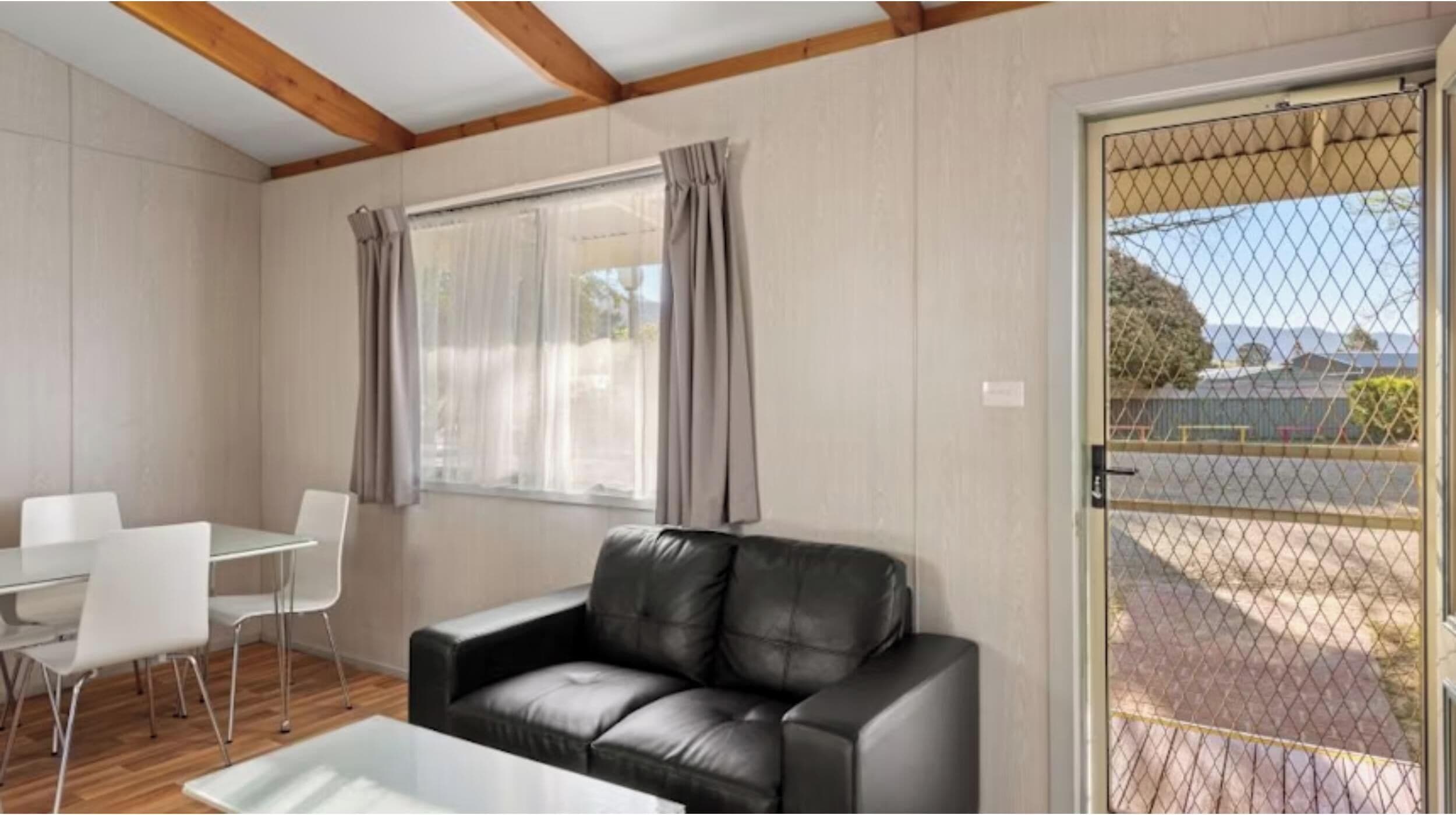 Una habitación con un sofá de cuero negro, sillas blancas, una mesita y una ventana con cortinas.