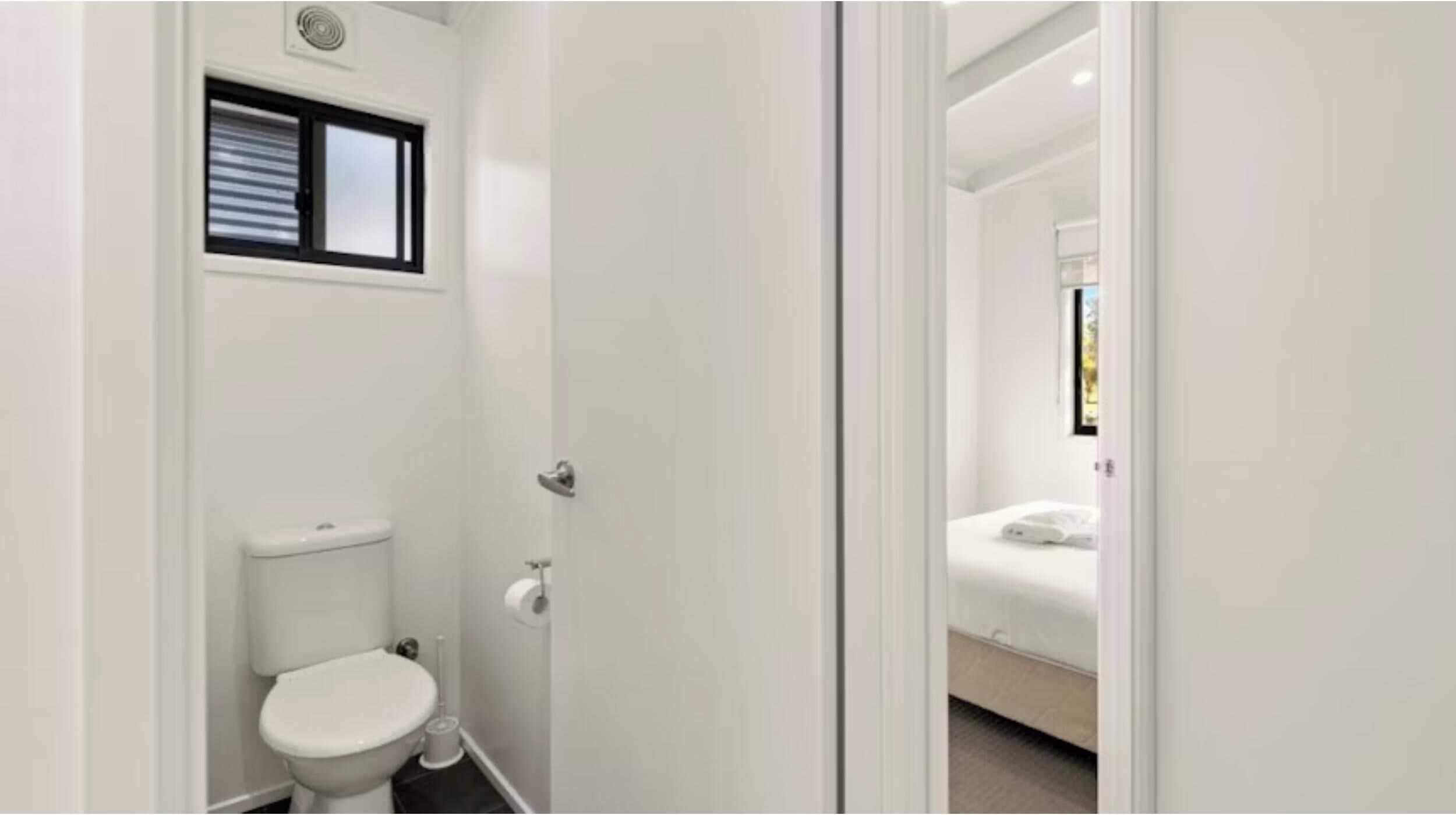 Un bagno piccolo e bianco con il water, una finestra e un ingresso che conduce a una camera da letto.