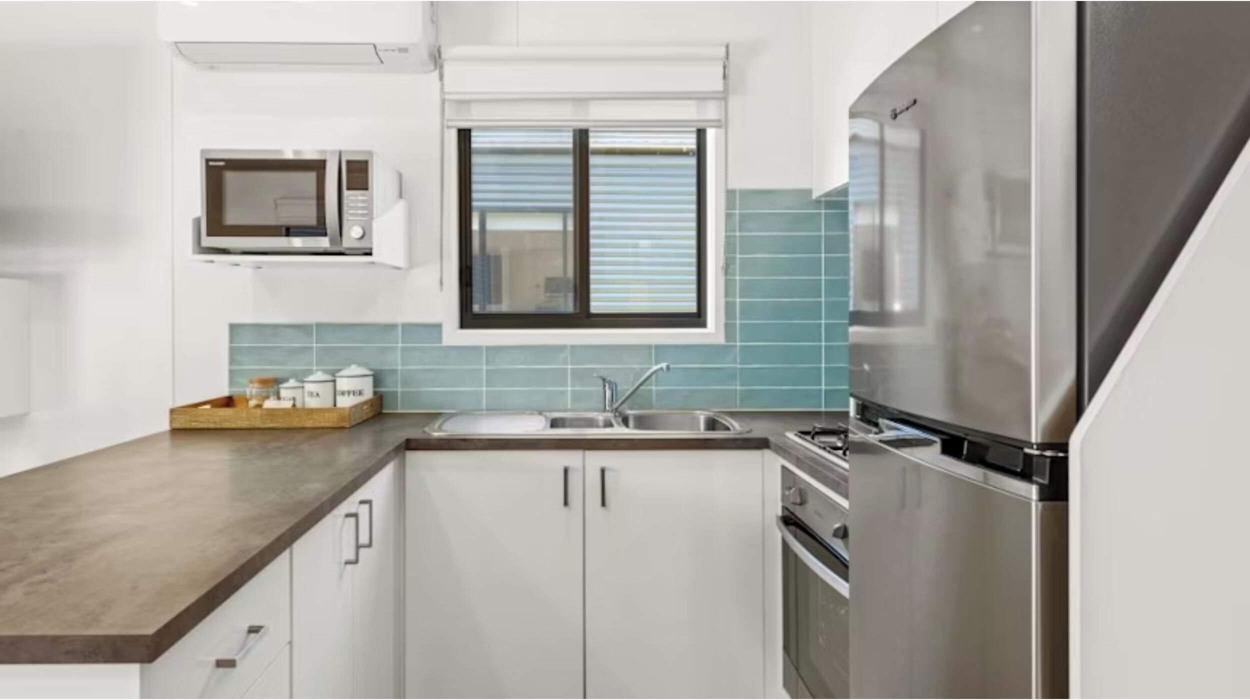 A modern kitchen with white cabinets, a stainless steel refrigerator, a microwave, and a window with blinds.