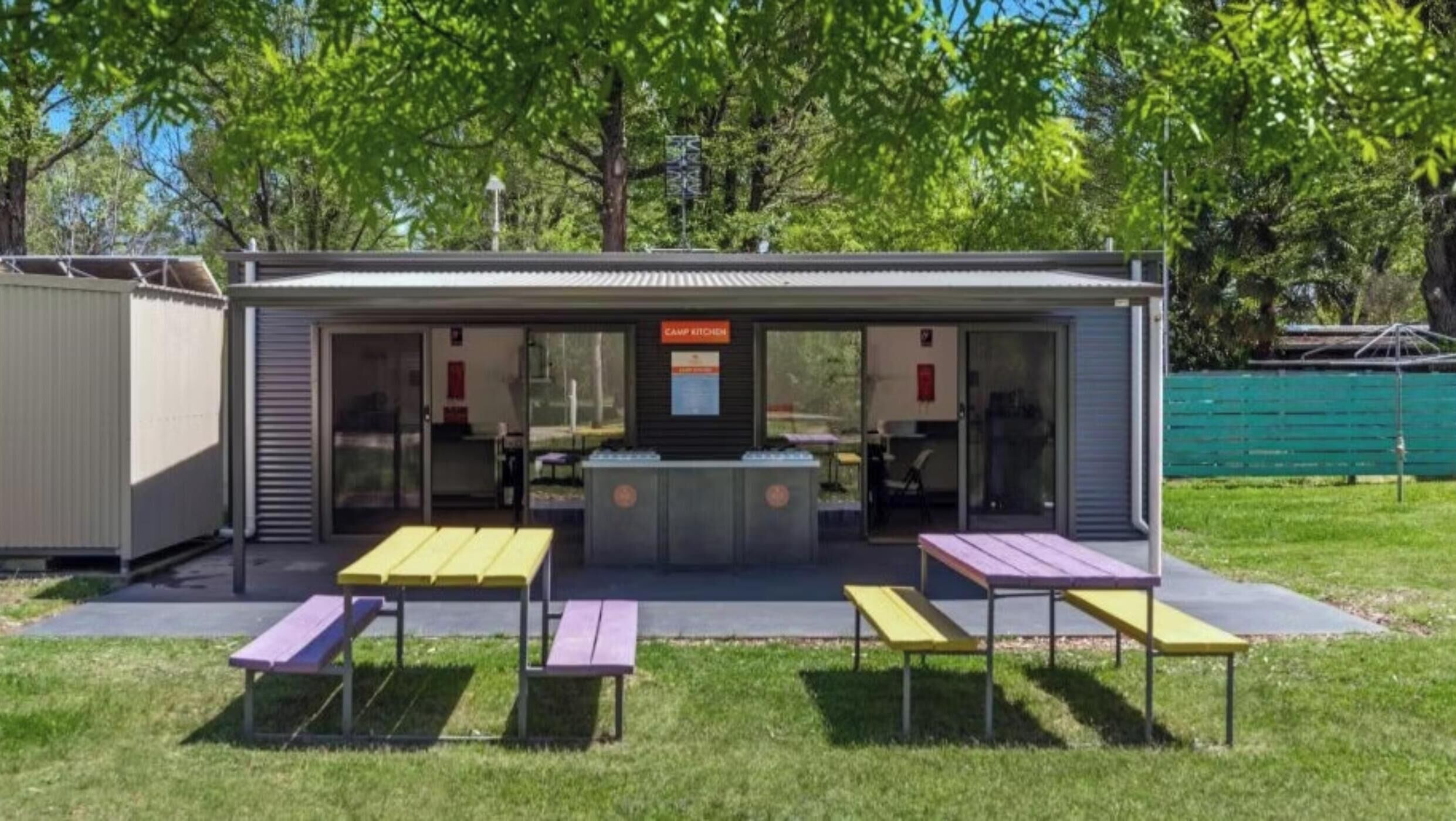 A small outdoor structure with a sign reading "CAMP KITCHEN," featuring a counter and seating area with tables and benches.