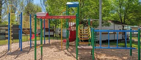 Un terrain de jeux doté de diverses installations, notamment un toboggan et des structures d’escalade, entouré d’arbres et comprenant un espace abrité.