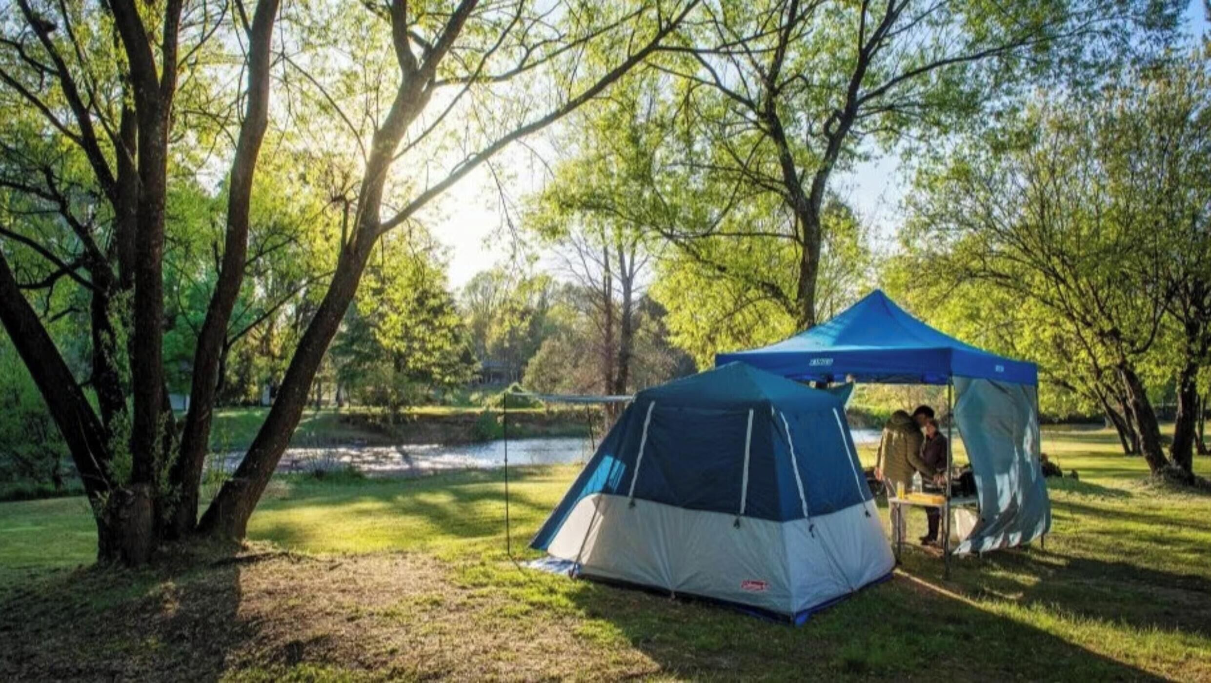 A blue and gray tent set up in a grassy area with trees and a body of water in the background.