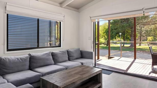 A modern living room with a grey sofa, a wooden coffee table, and a sliding glass door leading to an outdoor deck. - Discovery Parks - Mount Buffalo (Porepunkah)