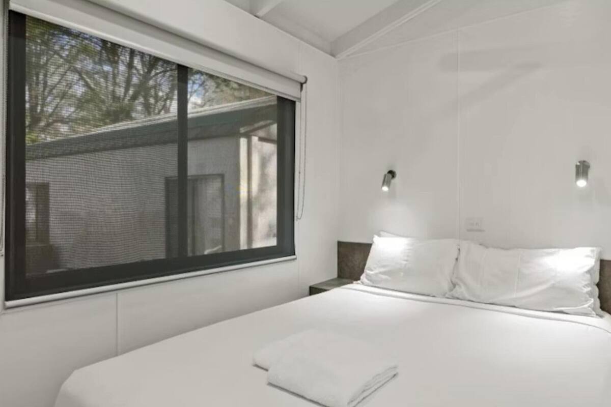 Ein Schlafzimmer mit einem großen Fenster, einem Bett mit weißer Bettwäsche, zwei Wandleuchten und einem Nachttisch.