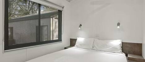 A bedroom with a large window, a bed with white linens, two wall-mounted lights, and a nightstand.