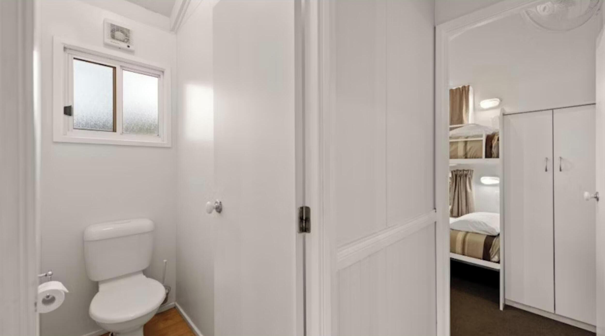 A bathroom with a toilet, a window, and a door leading to a bedroom with a bed and wardrobe.