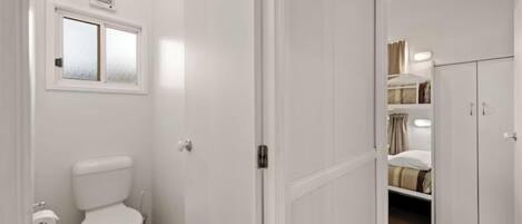 A bathroom with a toilet, a window, and a door leading to a bedroom with a bed and wardrobe.