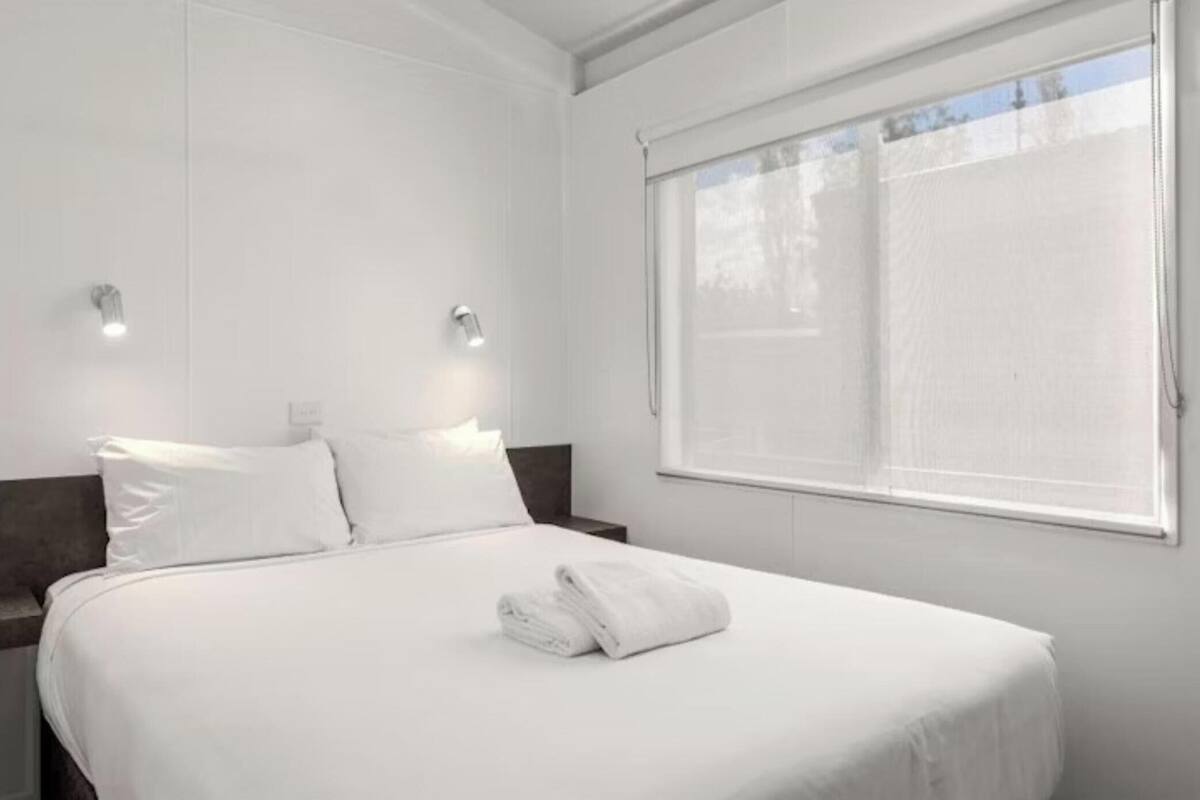 Ein Hotelzimmer mit einem großen Bett, zwei Nachttischen, einem Fenster mit Jalousien und einer Wandleuchte.