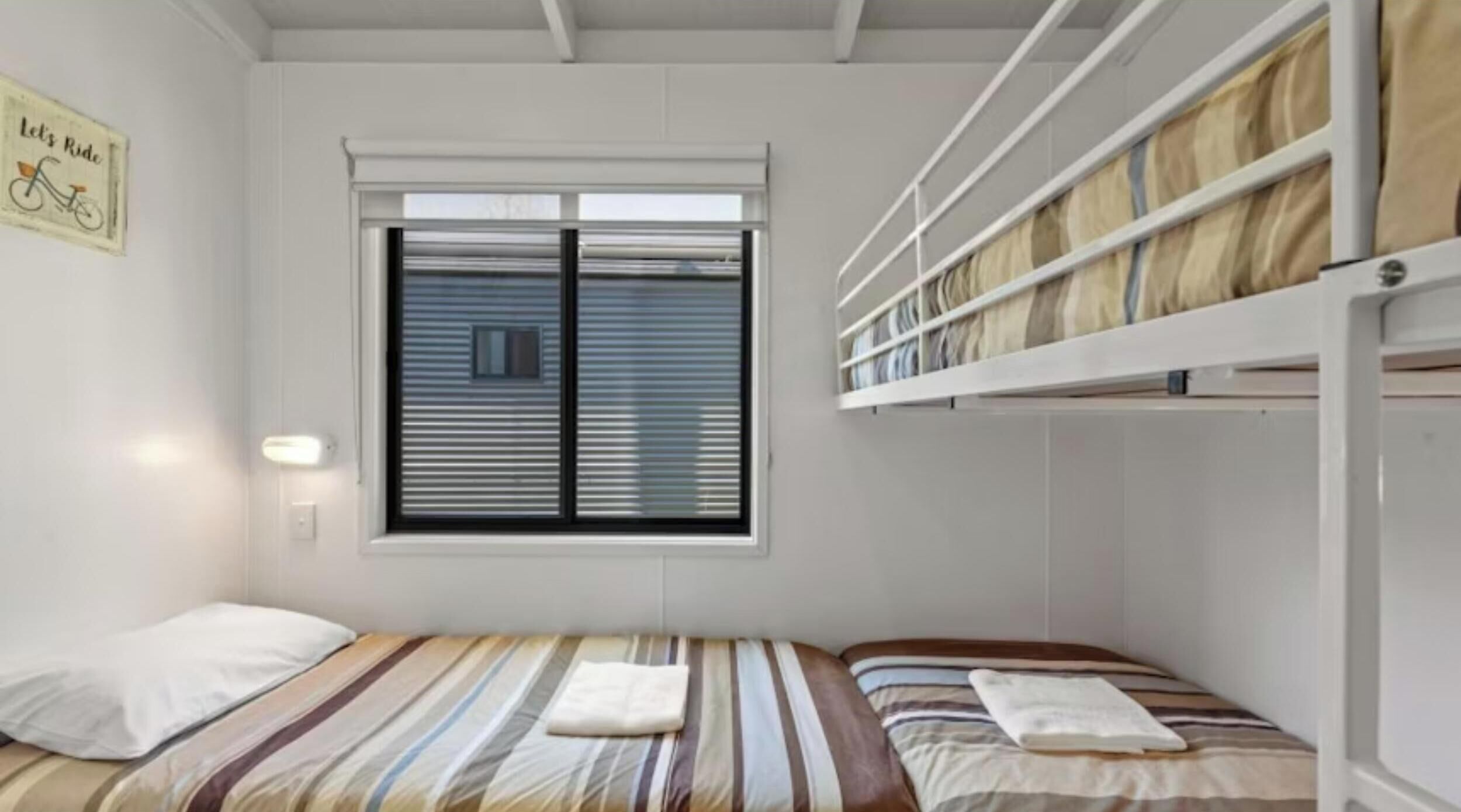 A small, single-room hotel room with a bunk bed, a window with blinds, a wall-mounted light, and a sign on the wall.