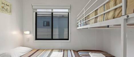 A small, single-room hotel room with a bunk bed, a window with blinds, a wall-mounted light, and a sign on the wall.