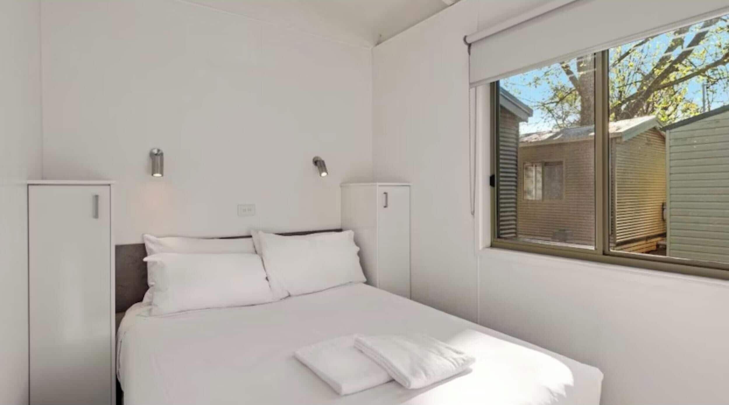 A bedroom with a bed, two bedside tables, a window, and a view of a house outside.