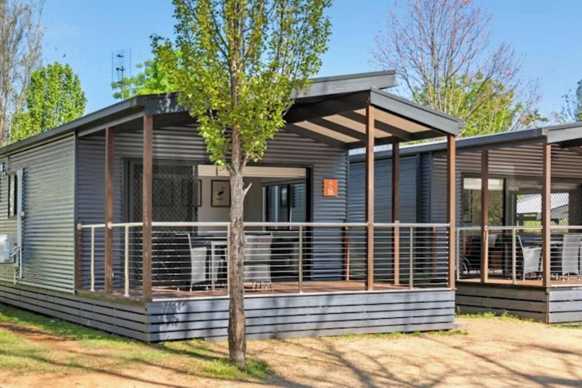Ein modernes, eingeschossiges Mobilheim mit überdachter Veranda und einem kleinen Baum davor.