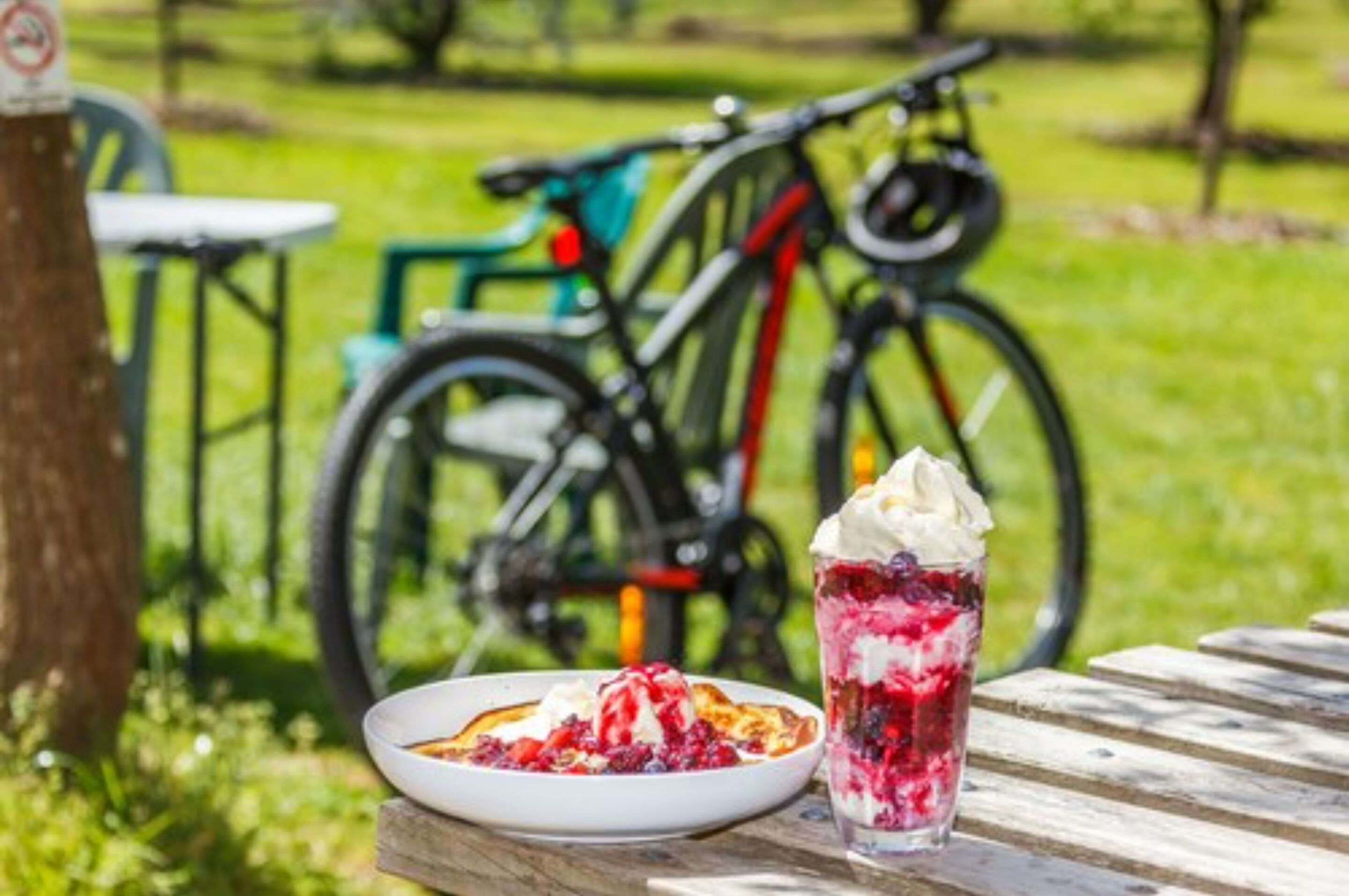 A picnic table with a plate of food and a glass of dessert in a park with bicycles in the background.