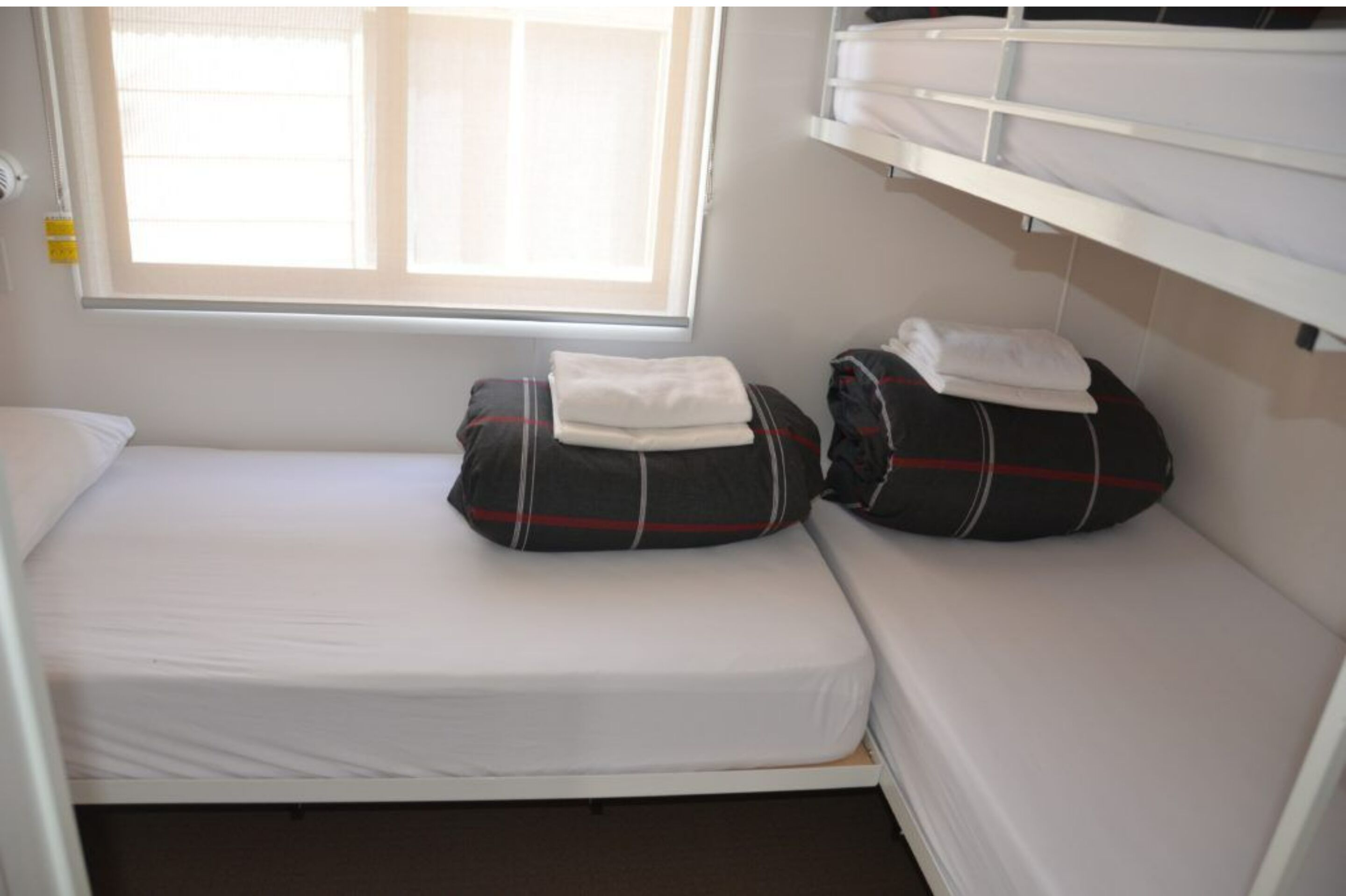 Une petite chambre avec un lit simple, une fenêtre laissant entrer la lumière naturelle, une tête de lit blanche et une étagère au-dessus du lit.