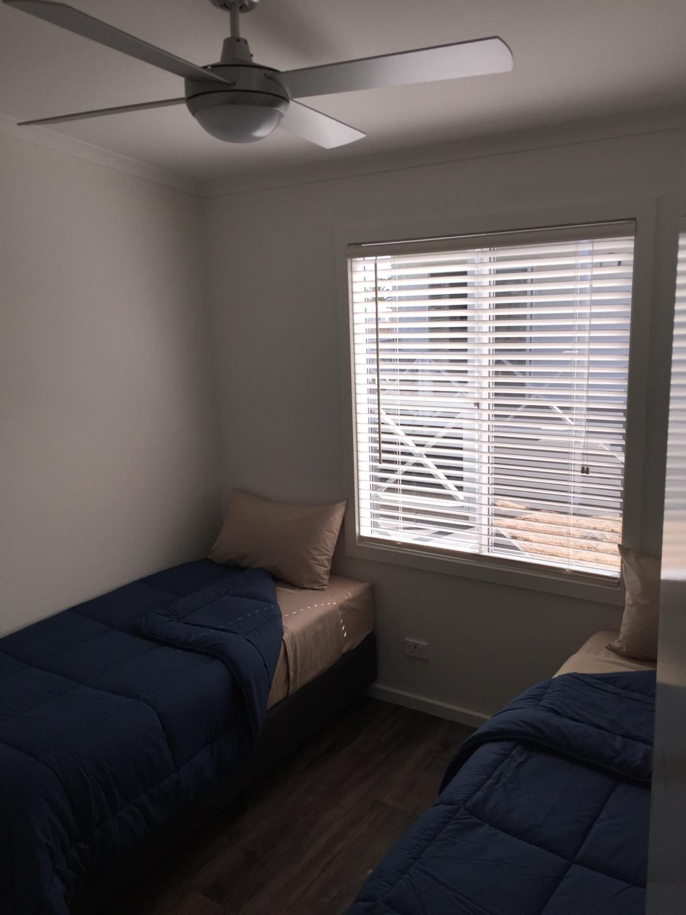 Une petite chambre avec un seul lit, dotée d’un ventilateur de plafond, d’une fenêtre avec stores et de deux lits recouverts de draps bleus.