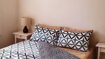A bed with a geometric patterned bedspread and pillows, a bedside table with a lamp, and a plain wall.