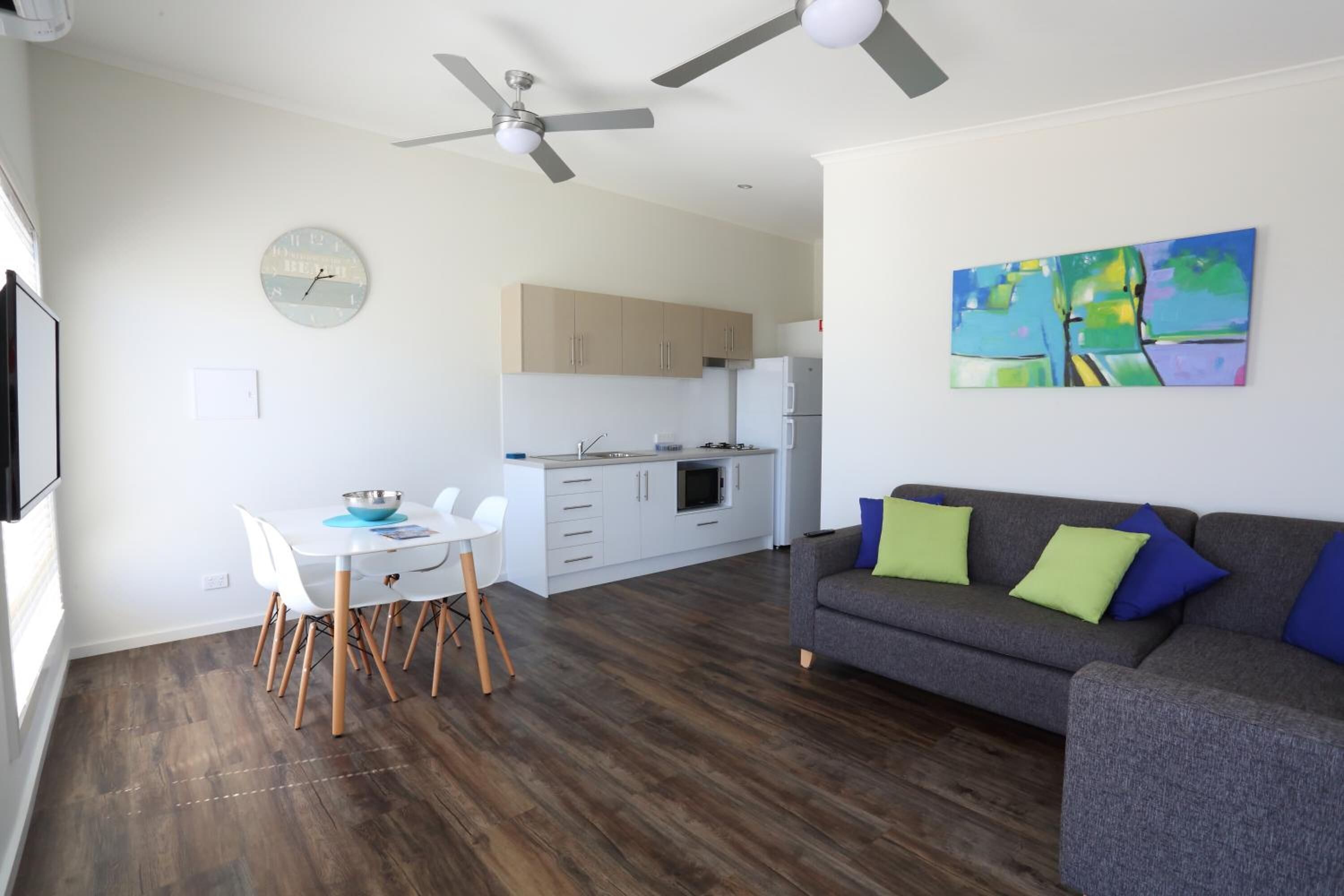 A modern living room with a gray sofa, a dining table with chairs, a kitchen area with cabinets and appliances, and a ceiling fan.