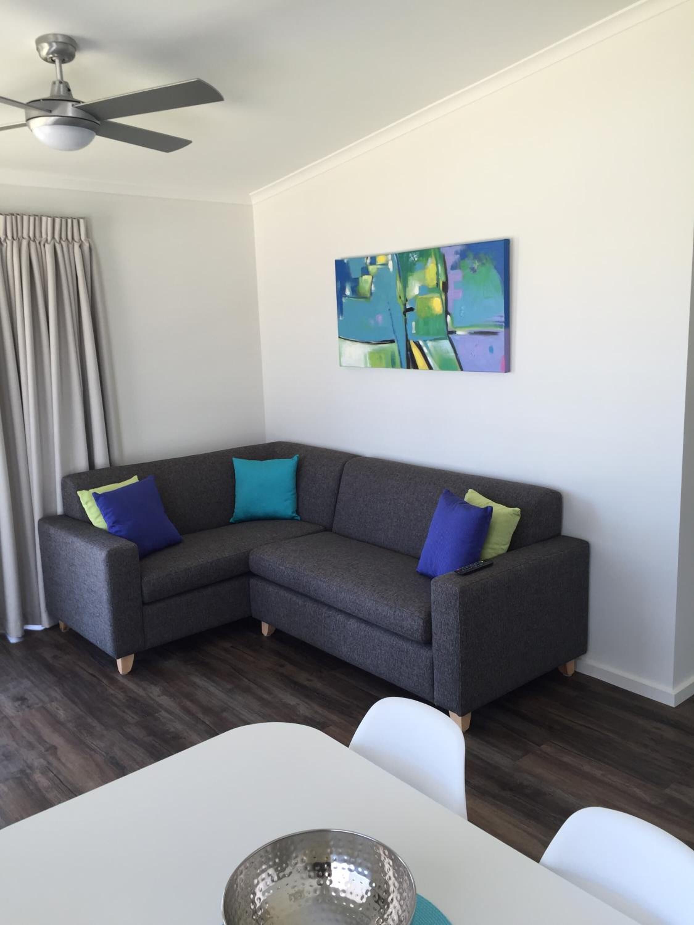 Un salon moderne avec un canapé d’angle gris, un ventilateur de plafond, un tableau et une table à manger avec des chaises.