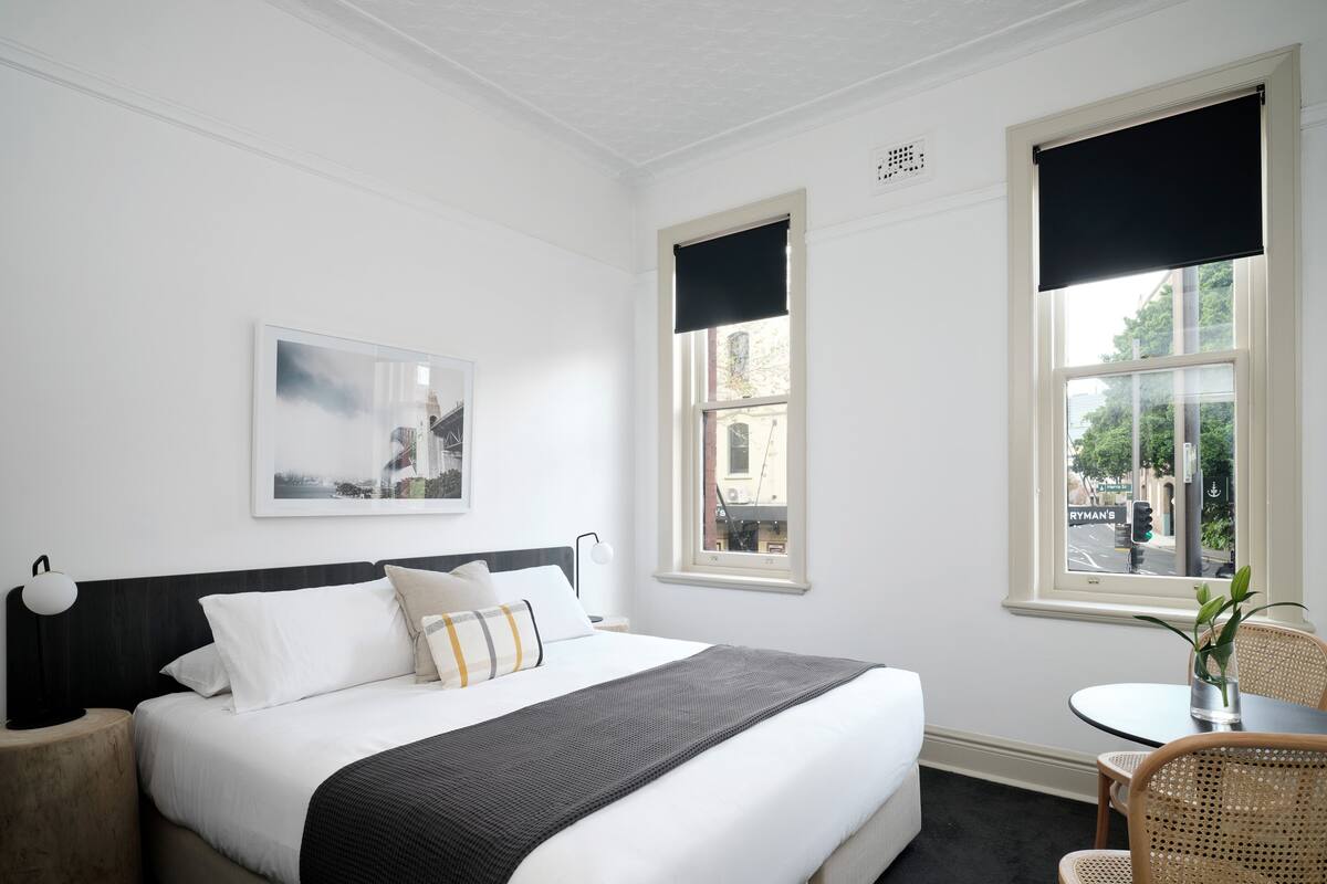 A hotel room with a bed, a bedside table, a chair, a window with a view of the street, and a framed picture on the wall.