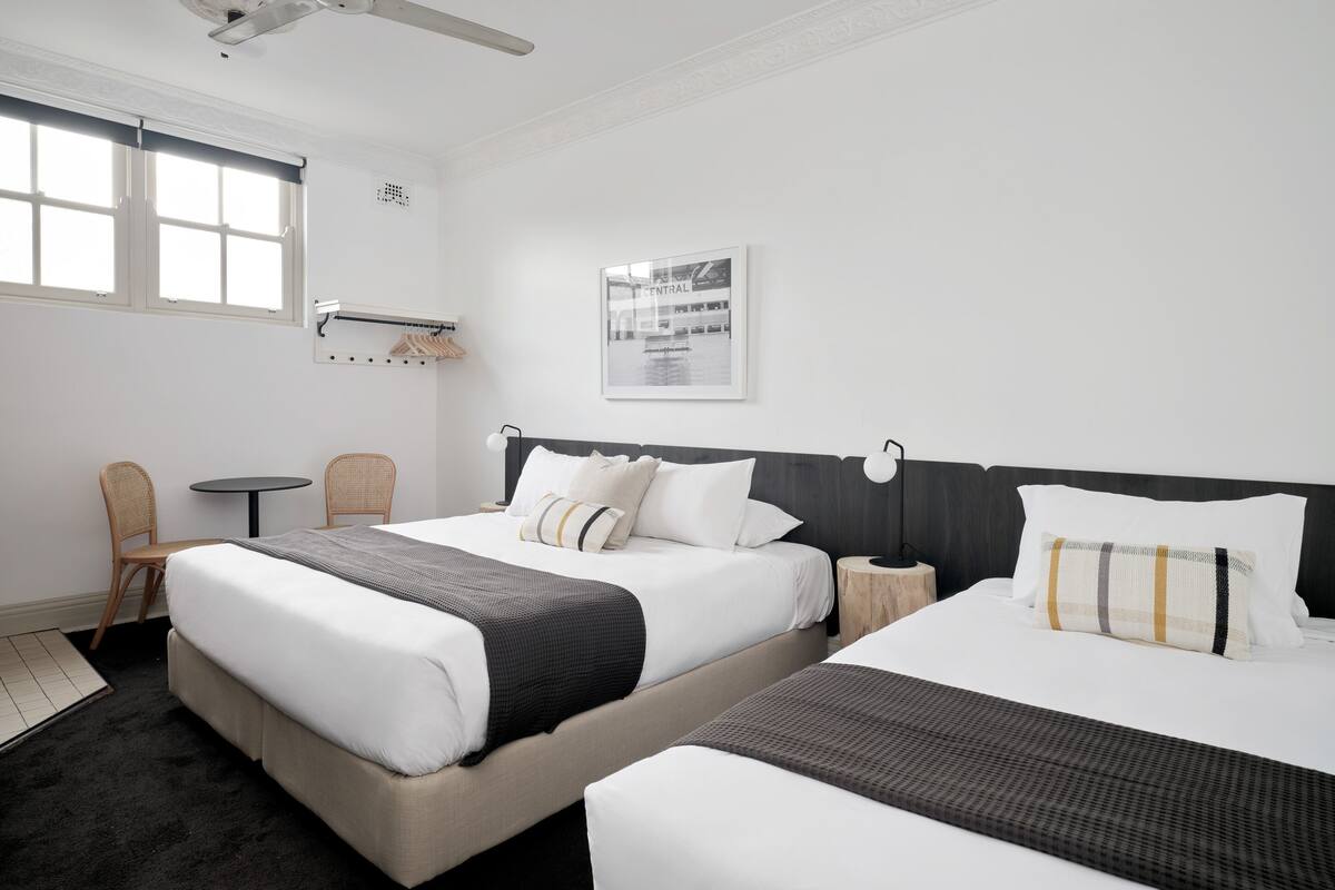 A hotel room with two beds, a ceiling fan, a small table, two chairs, a window, and a wall-mounted shelf with clothes.