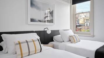 Habitación de hotel con dos camas, un cuadro enmarco con una vista urbana, una mesita de noche y una ventana que ofrece vistas de edificios.