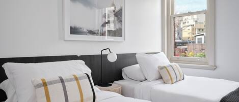 Habitación de hotel con dos camas, un cuadro enmarco con una vista urbana, una mesita de noche y una ventana que ofrece vistas de edificios.