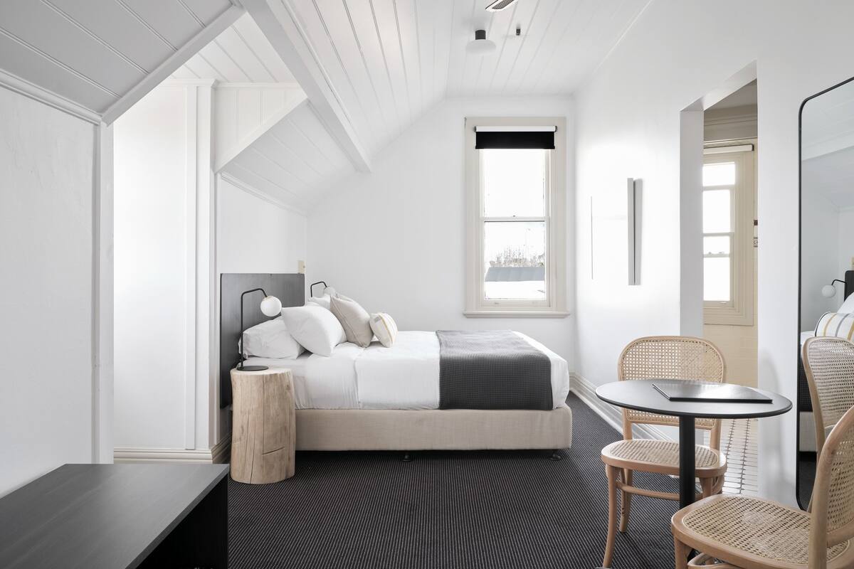 A modern hotel room with a bed, bedside table, chair, and a small table with a chair.