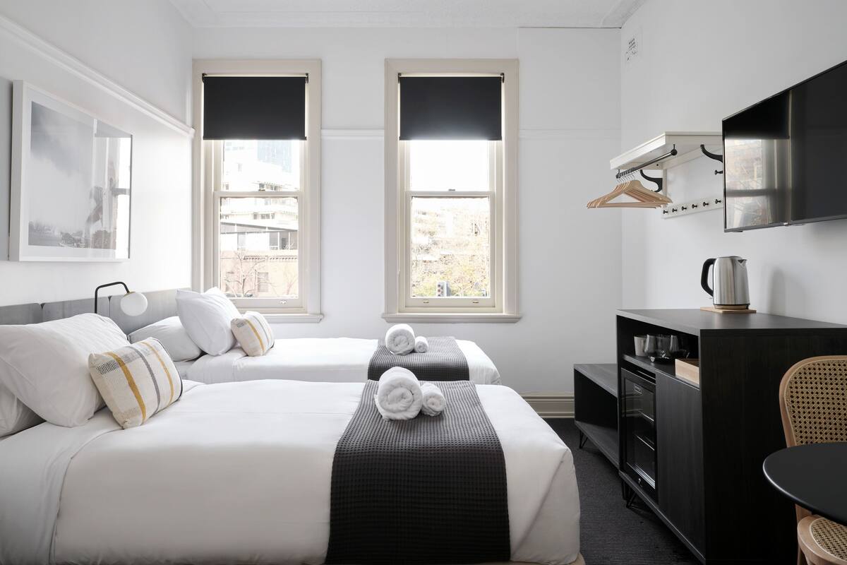 A hotel room with two beds, a desk, a chair, a TV, and a window with a view of buildings.