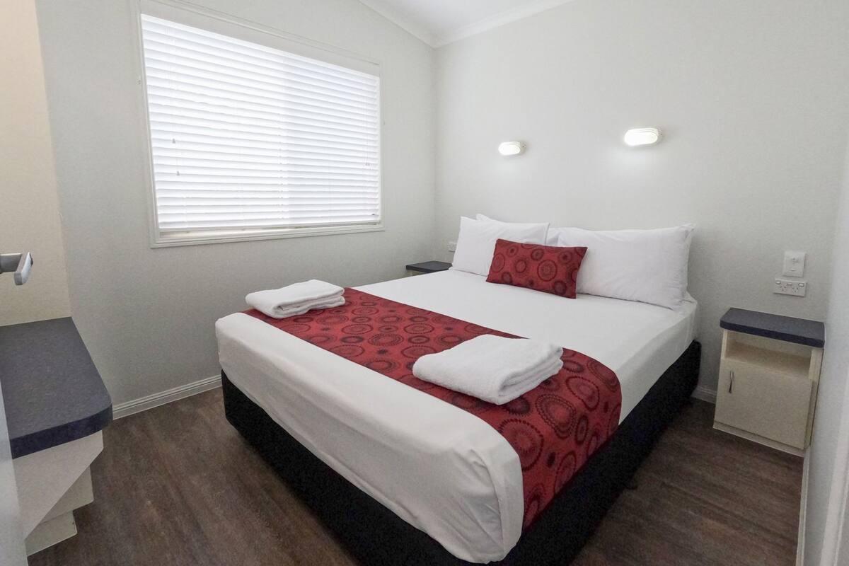 A hotel room with a bed, a window with blinds, a bedside table, and a nightstand.