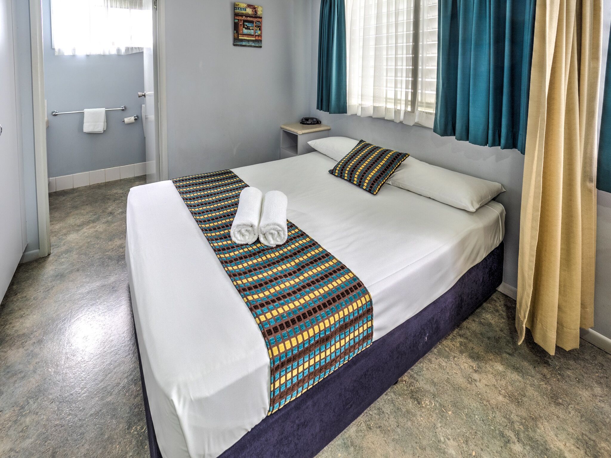 A hotel room with a bed, a bedside table, a window with curtains, and a bathroom visible in the background.