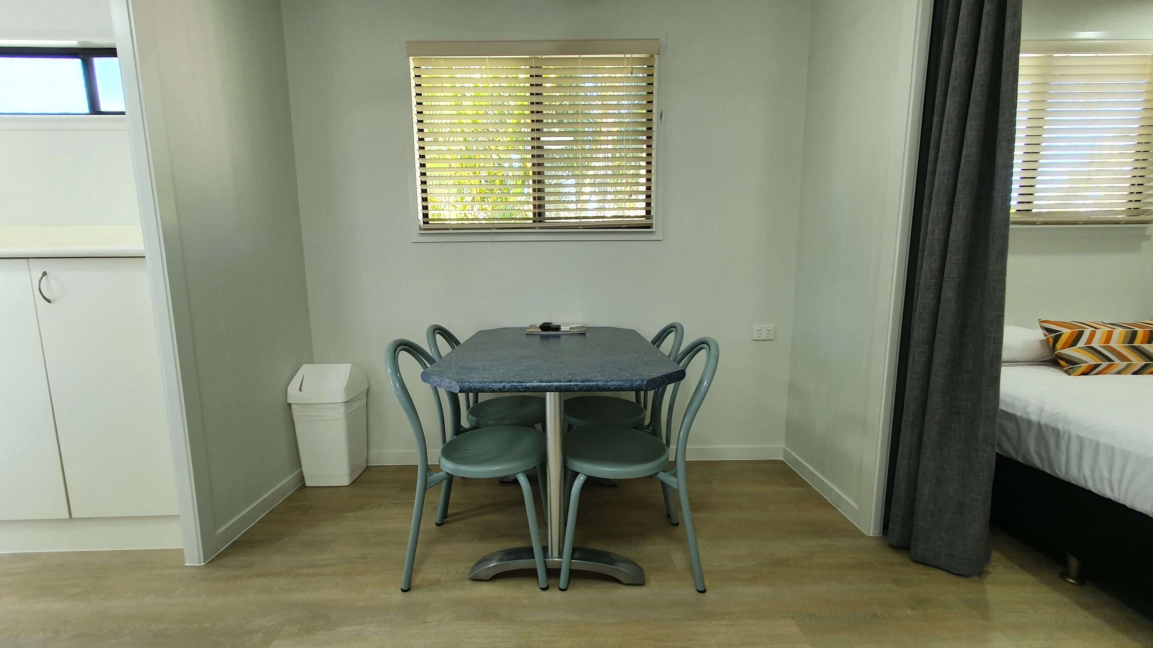 A compact room with a dining table and chairs, a window with blinds, a bed with a patterned pillow, and a small trash bin.