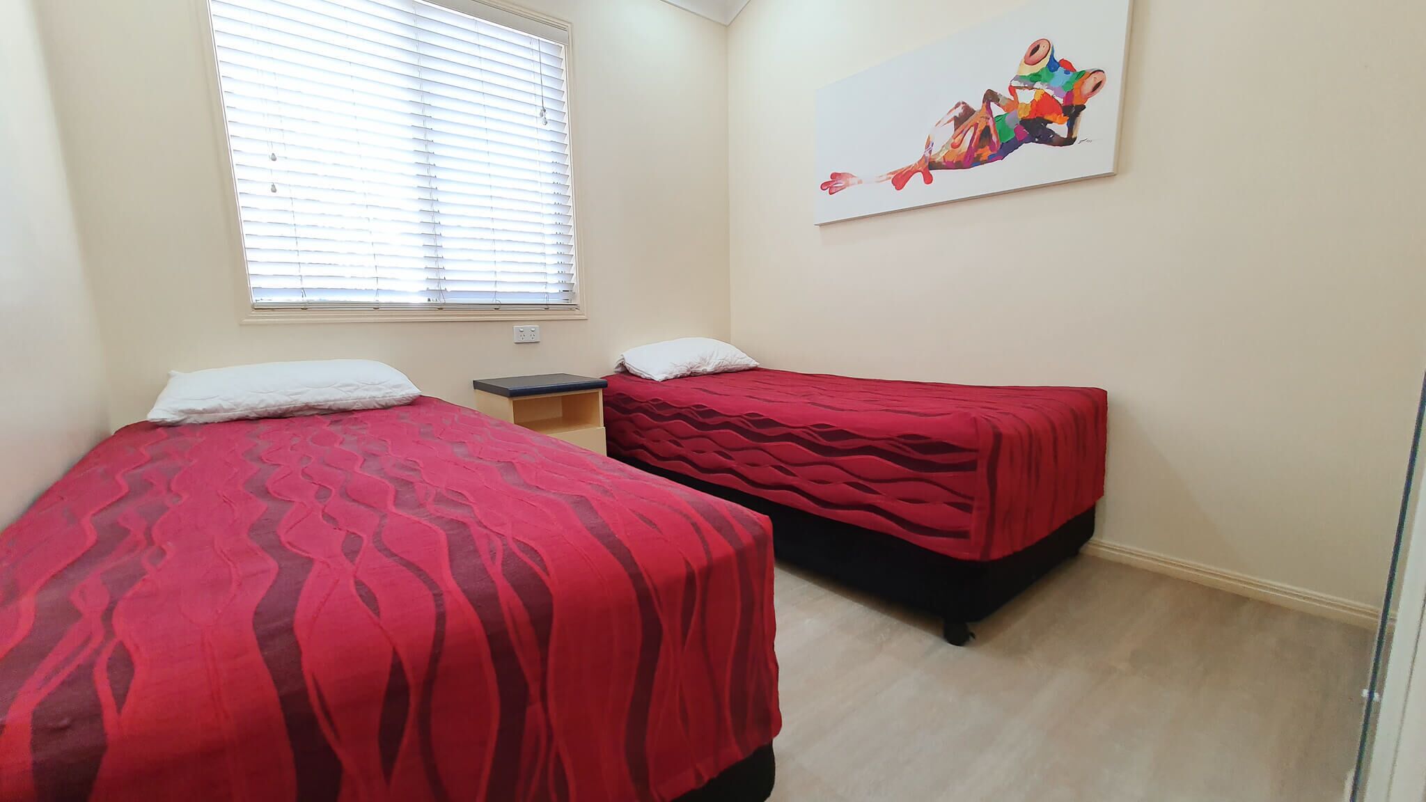 A room with two beds, each with a red bedspread, a small bedside table, and a wall-mounted painting.