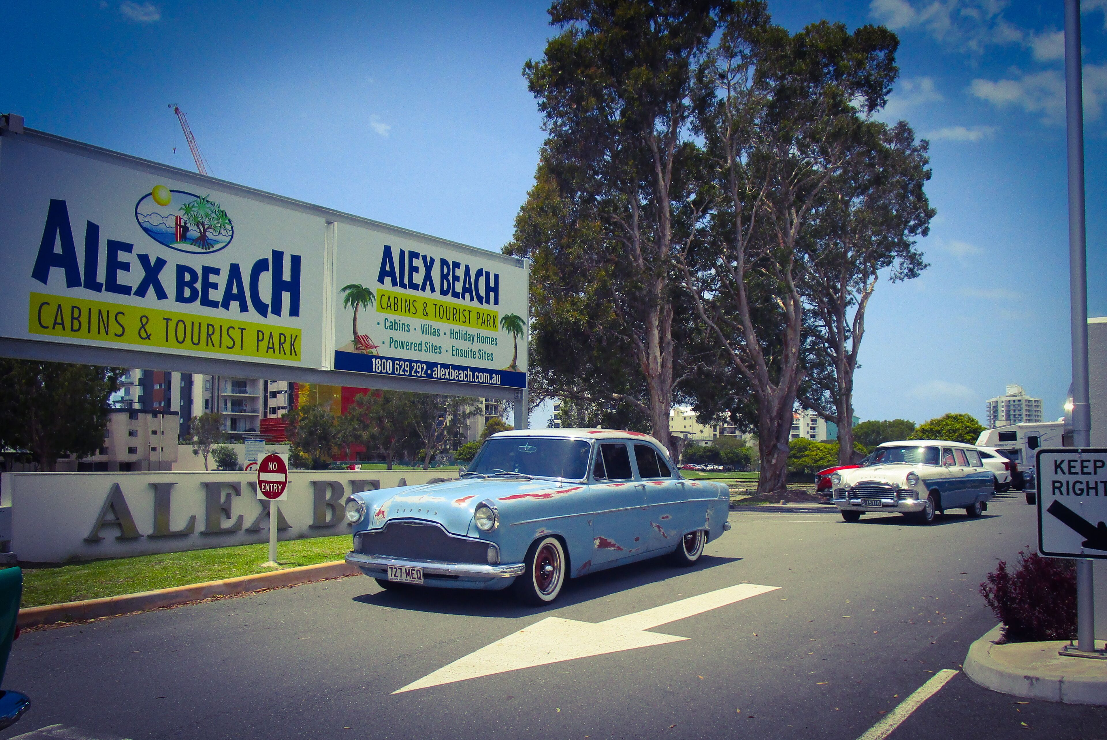 A vintage car parked in front of a building with a sign for Alex Beach, featuring cabins and tourist park amenities.