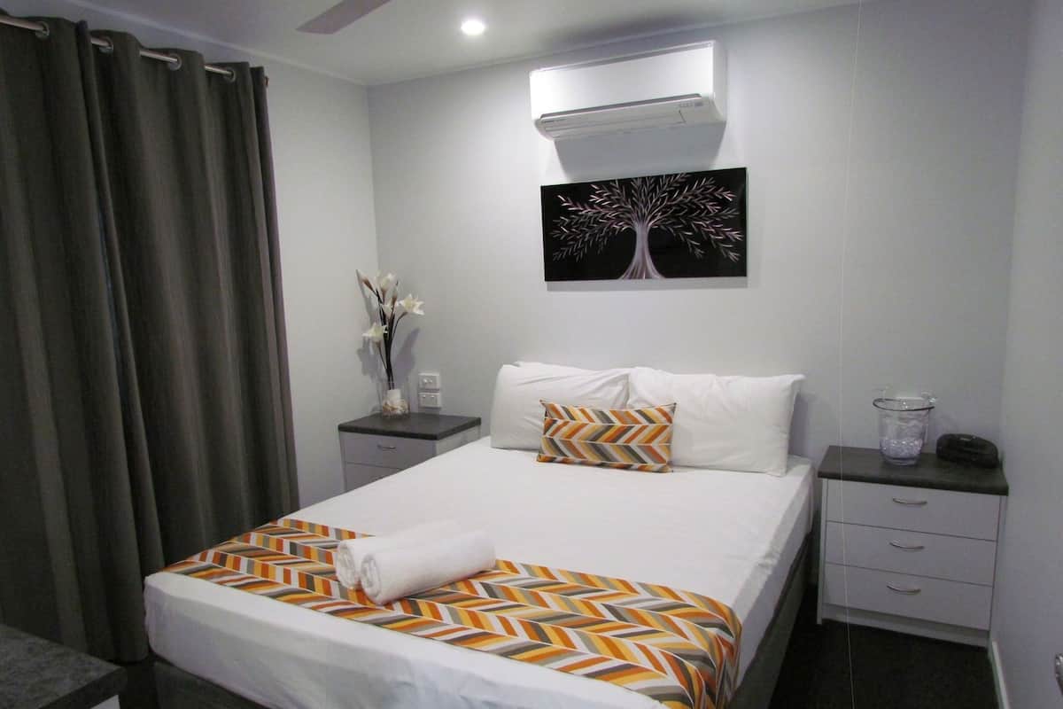 A hotel room with a bed, a nightstand, a wall-mounted air conditioner, and a painting of a tree.