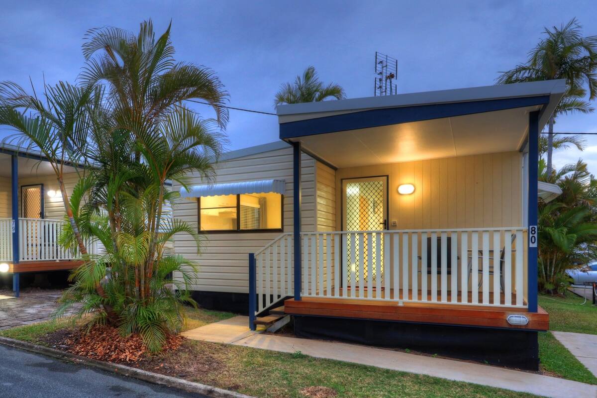 A row of mobile homes with wooden decks and railings, surrounded by palm trees.