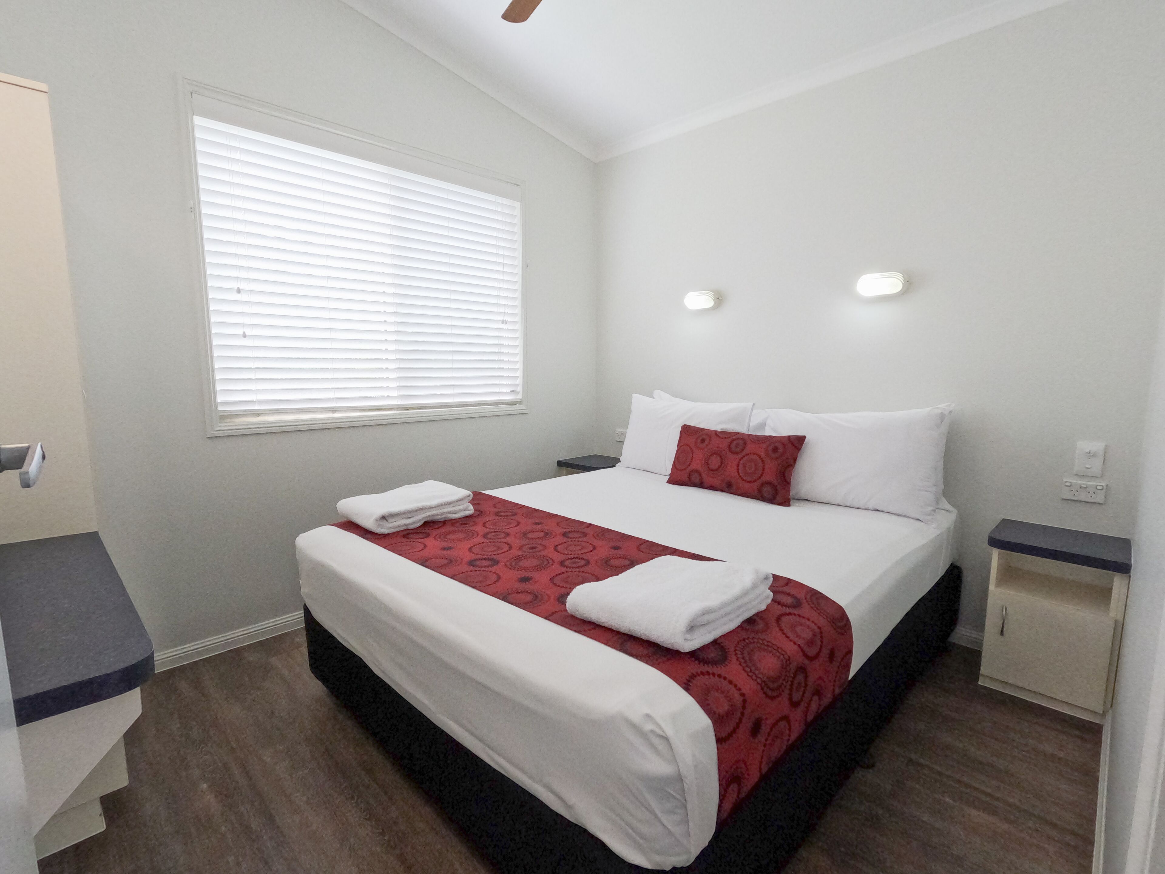 Uma cama bem arrumada com roupa de cama branca e vermelha, uma janela com persianas, uma mesa de cabeceira e um aplique de parede.