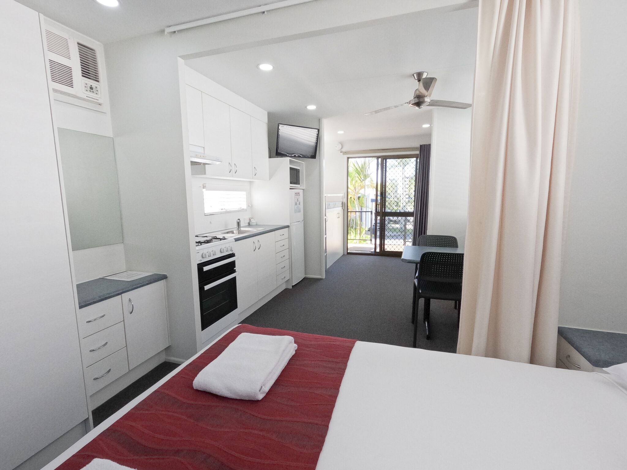 A modern hotel room with a kitchenette, a bed with a red accent, a TV, and a balcony with curtains.