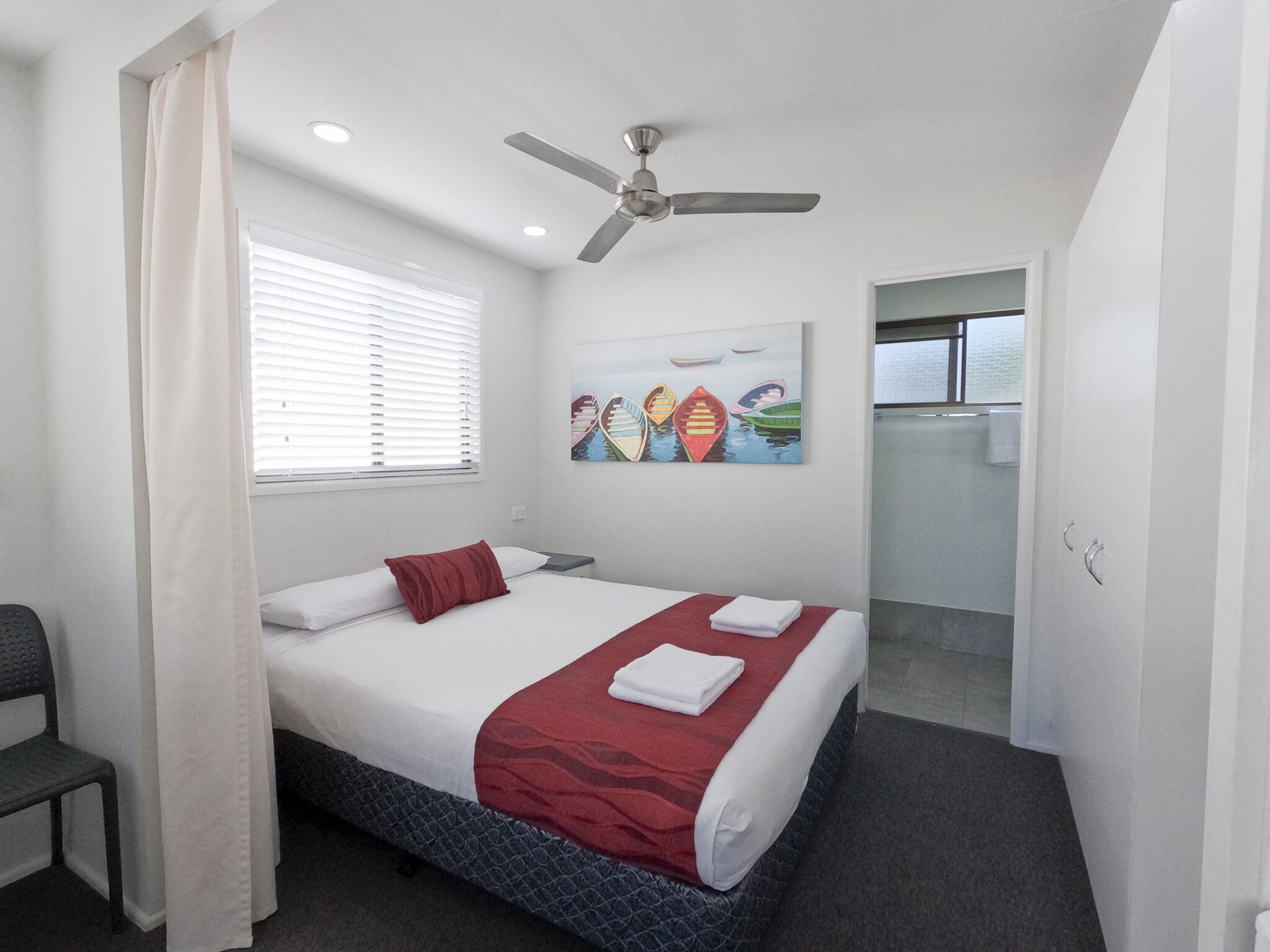 Una camera d'albergo con un letto, una ventola a soffitto, una finestra con persiane, un quadro raffigurante barche e un bagno visibile attraverso una porta aperta.