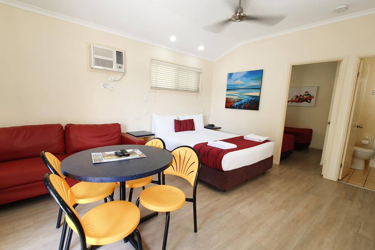 A hotel room with a bed, a red sofa, a dining table with chairs, a ceiling fan, and a painting on the wall.