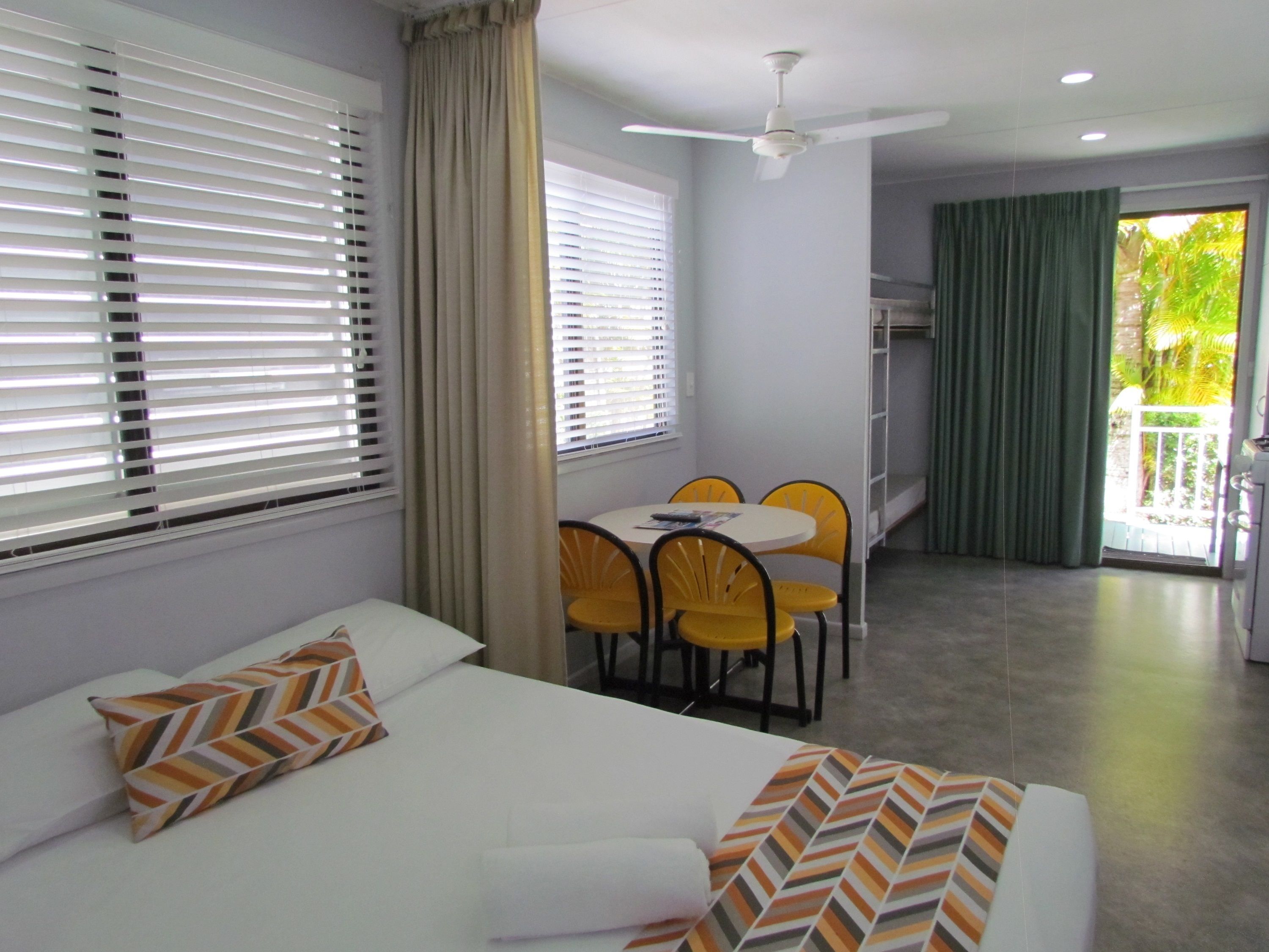 Habitación de hotel con cama, mesa de comedor con sillas, ventilador de techo y vistas al exterior.