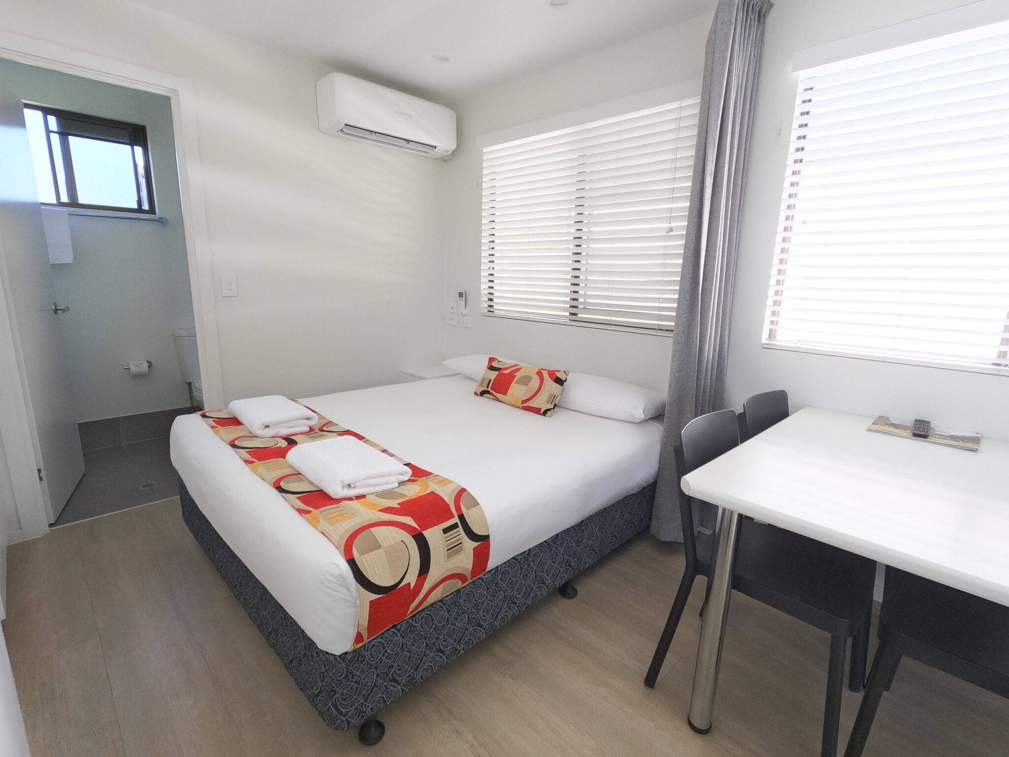 Una cama individual con una colcha estampada, una mesita de noche, una silla, una ventana con persianas y un equipo de aire acondicionado.