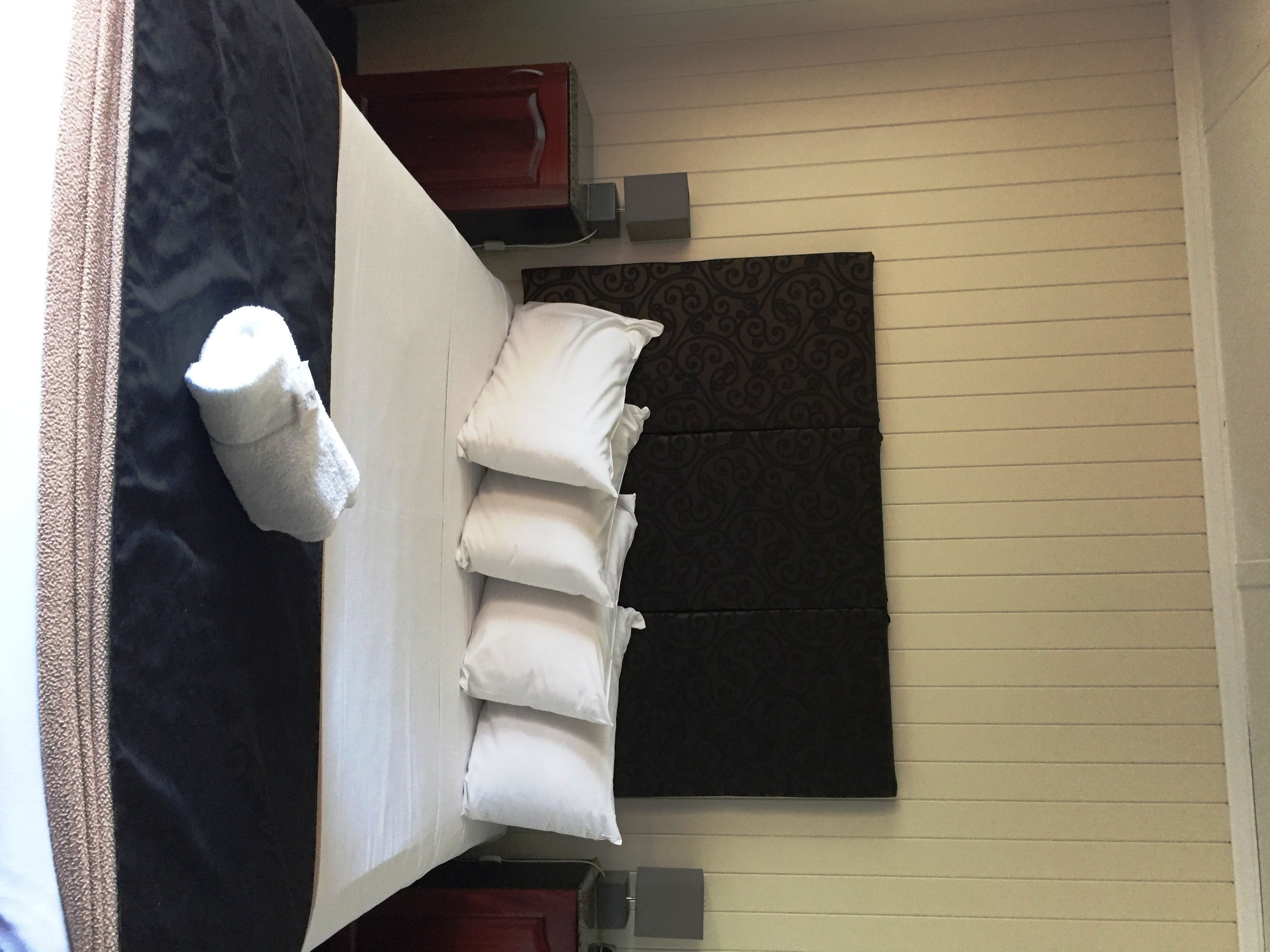 A hotel room with a bed, pillows, a blanket, a dark headboard, a patterned wall, and a red cabinet.