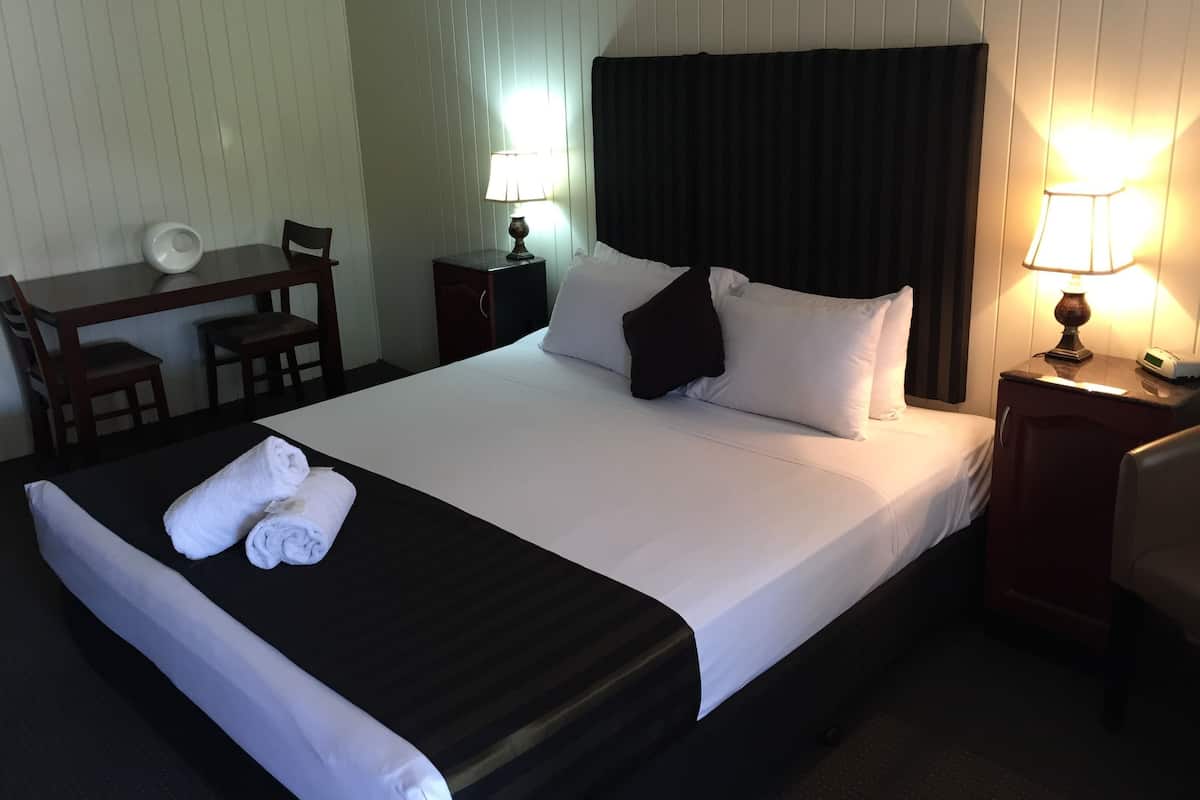 A neatly made bed with white linens, a black headboard, and a black pillow. There is a desk with a chair, a small table with a lamp, and a wall-mounted clock.